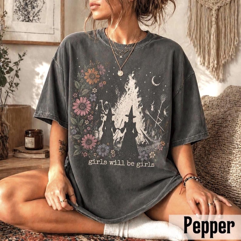 May include: Dark gray t-shirt featuring a floral and witch-themed graphic. The design includes two witch silhouettes, a bonfire, a crescent moon, and the text "girls will be girls". The shirt has a vintage, worn look.