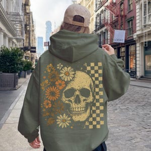 May include: An olive green hoodie featuring a skull design with floral and checkered elements. The skull is in the center, with flowers on the left and a checkered pattern on the right. The hoodie has a hood and long sleeves.