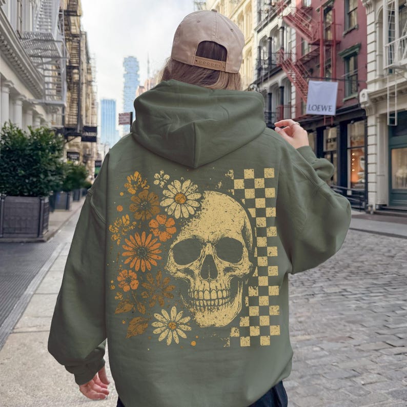 May include: An olive green hoodie featuring a skull design with floral and checkered elements. The skull is in the center, with flowers on the left and a checkered pattern on the right. The hoodie has a hood and long sleeves.