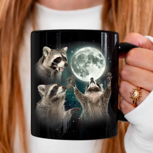 May include: Black ceramic mug featuring three raccoons with a full moon and starry background. Two raccoons look up at the moon, while the third appears to be howling. The mug has a black handle.