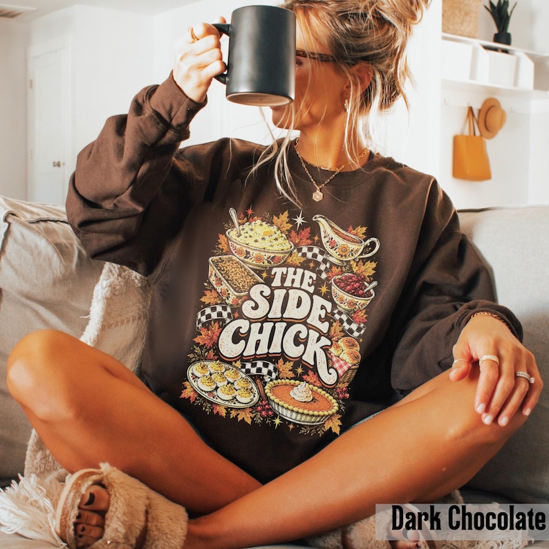 May include: A dark chocolate brown long-sleeve sweatshirt featuring a colorful graphic print of various Thanksgiving side dishes, including a pie and casserole, with the text "THE SIDE CHICK" in a bold, retro-inspired white and orange font.