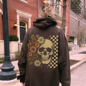 May include: A brown hoodie featuring a skull design with floral and checkered accents. The skull is beige, and the flowers are in shades of orange and yellow. The hoodie has a hood and long sleeves.