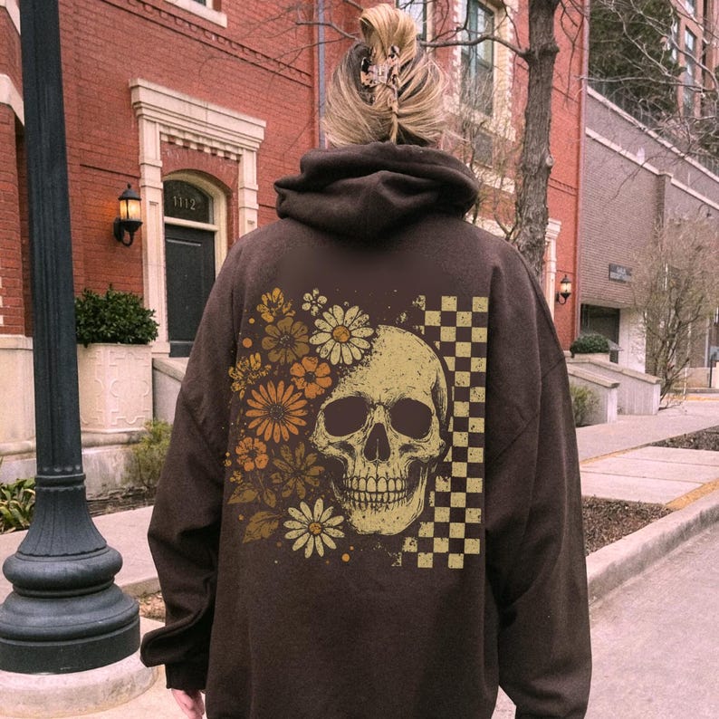 May include: A brown hoodie featuring a skull design with floral and checkered accents. The skull is beige, and the flowers are in shades of orange and yellow. The hoodie has a hood and long sleeves.