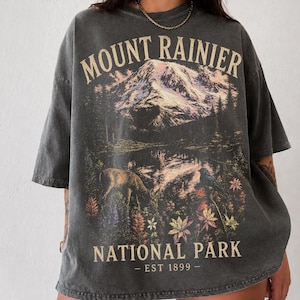 以下が含まれることがあります： マウントレーニア国立公園のデザインが施された濃いグレーのTシャツ。グラフィックには山並み、鹿、花が含まれています。シャツのテキストは「MOUNT RAINIER NATIONAL PARK - EST 1899 - 」です。