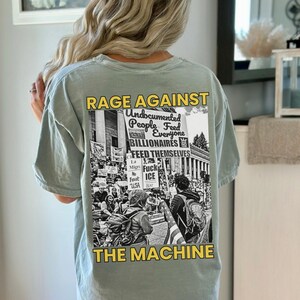 Puede incluir: Camiseta verde claro con un gr&aacute;fico en la espalda. El gr&aacute;fico es una imagen en blanco y negro de una protesta con las palabras "RAGE AGAINST THE MACHINE" y otro texto. La camiseta es de estilo casual.