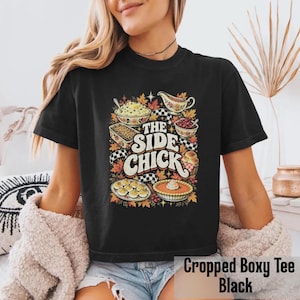 May include: A black short-sleeve t-shirt featuring a graphic print of various Thanksgiving side dishes, including mashed potatoes, stuffing, and pie. The text "THE SIDE CHICK" is printed in large, bold, cream-colored lettering in the center.