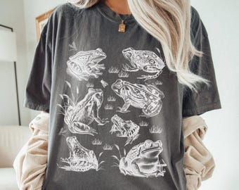 May include: Dark grey t-shirt with a white illustration of multiple frogs in different positions. The design shows detailed drawings of the amphibians, some with grass and insects. The shirt has a relaxed fit and short sleeves.