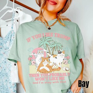 May include: A light green t-shirt with a graphic print of cats on a beach with palm trees. The text on the shirt reads "If you like Trump then you probably won't like me and I'm okay with that".