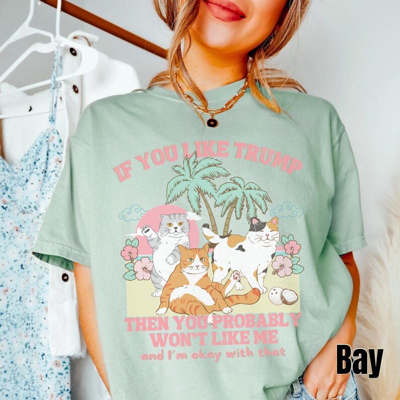 May include: A light green t-shirt with a graphic print of cats on a beach with palm trees. The text on the shirt reads "If you like Trump then you probably won't like me and I'm okay with that".