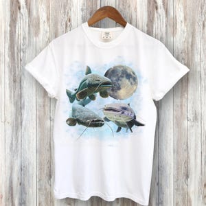 May include: White t-shirt with a graphic print of three catfish swimming under a full moon. The catfish are light blue and gray with white accents.