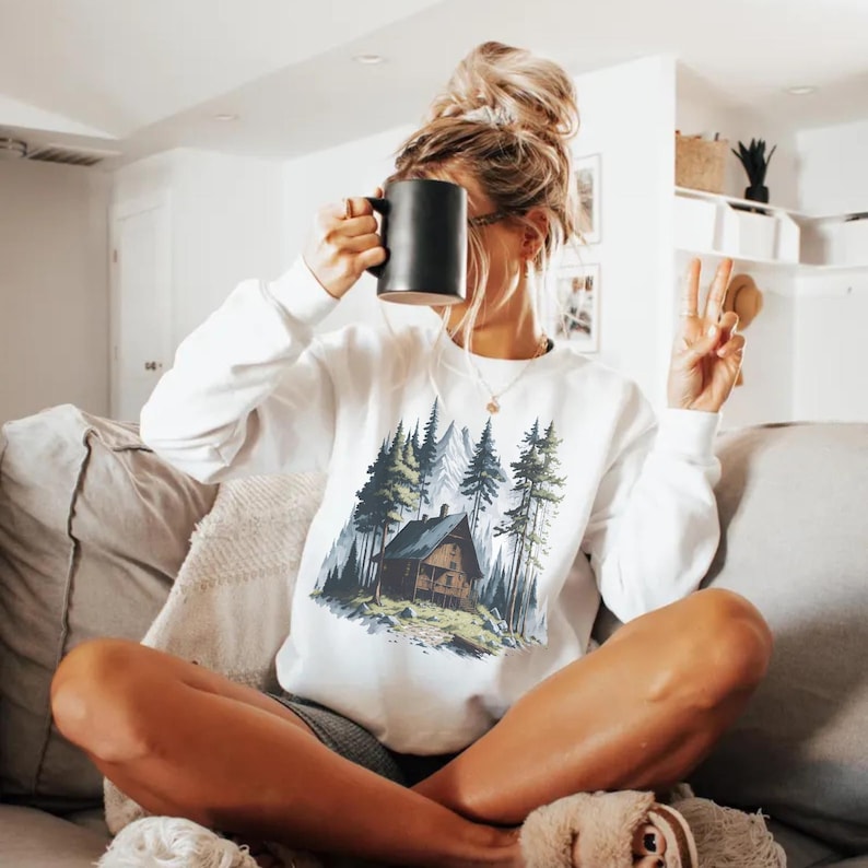 May include: White sweatshirt featuring a cabin scene with trees and mountains. The graphic has a rustic, outdoorsy theme. The sweatshirt is long-sleeved and has a crew neck. The person is holding a black mug.
