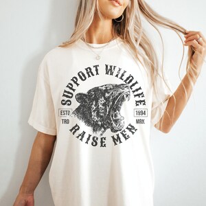 May include: A white t-shirt with a black graphic print of a roaring animal head. The graphic says "Support Wildlife" and "Raise Men" with the text "Est. 1994" in a small rectangle.