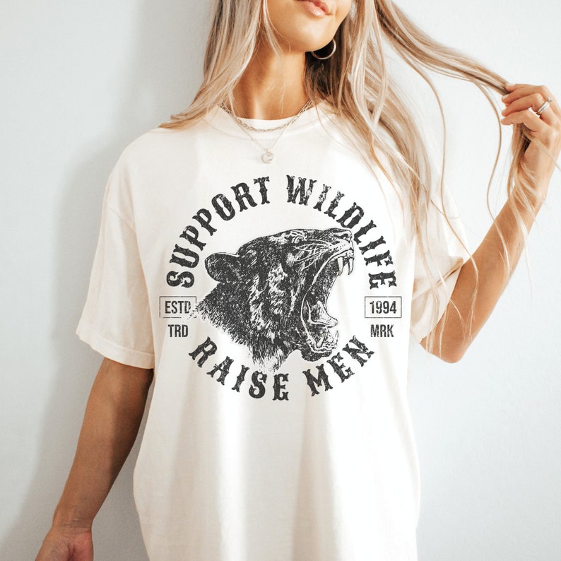 May include: A white t-shirt with a black graphic print of a roaring animal head. The graphic says "Support Wildlife" and "Raise Men" with the text "Est. 1994" in a small rectangle.