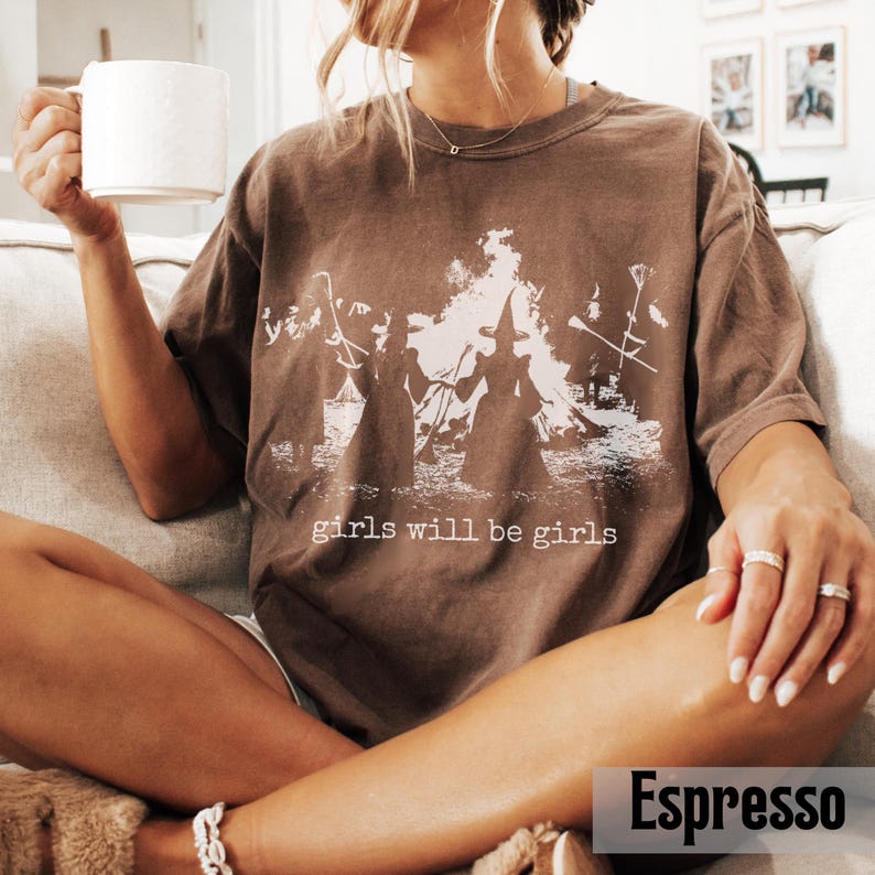 Pu&ograve; includere: Una t-shirt marrone a maniche corte con una stampa grafica bianca di due figure con cappelli a punta davanti a un grande fal&ograve; con scope e il testo "girls will be girls" stampato in un carattere serif bianco sul petto.
