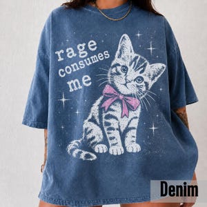 May include: A blue acid-wash t-shirt featuring a white graphic of a kitten wearing a pink bow. The text "rage consumes me" is printed in white above the kitten, surrounded by small white star illustrations across the front of the garment.