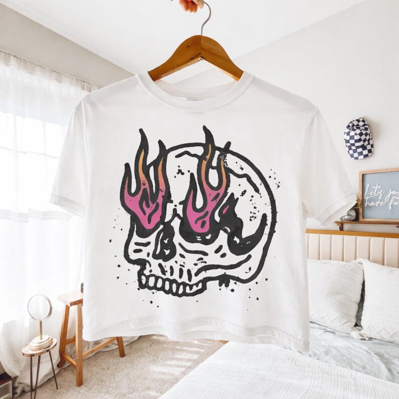 May include: White t-shirt featuring a black and pink skull graphic with flames emanating from the eye sockets. The design is a bold, graphic illustration. The shirt is hanging on a wooden hanger.