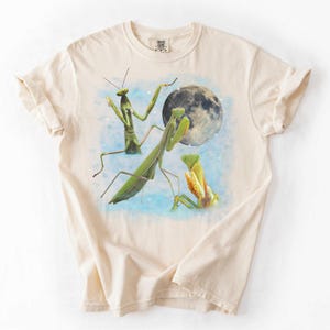 Puede incluir: Camiseta color crema con un dise&ntilde;o caprichoso de tres mantis religiosas y una luna llena. Las mantis son verdes y amarillas, sobre un cielo azul claro. Una camiseta gr&aacute;fica &uacute;nica.
