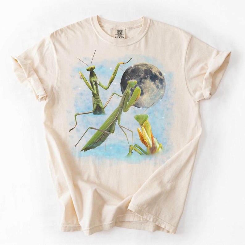 Puede incluir: Camiseta color crema con un dise&ntilde;o caprichoso de tres mantis religiosas y una luna llena. Las mantis son verdes y amarillas, sobre un cielo azul claro. Una camiseta gr&aacute;fica &uacute;nica.