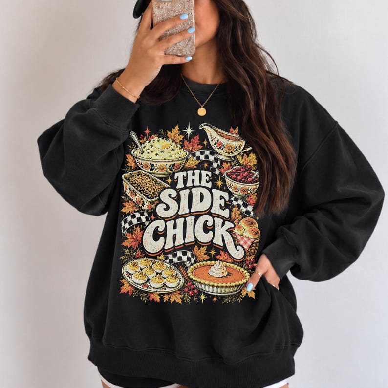 May include: A black crewneck sweatshirt featuring a graphic print with the text THE SIDE CHICK in large white letters. The design includes illustrations of various food dishes, autumn leaves, and a black and white checkered pattern border.