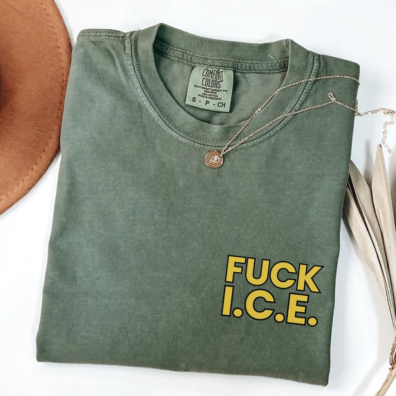 Puede incluir: Camiseta verde oliva con el texto "FUCK I.C.E." en letras amarillas. La camiseta tiene cuello redondo y una peque&ntilde;a etiqueta en el cuello. Un collar dorado con un colgante est&aacute; drapeado sobre la camiseta.