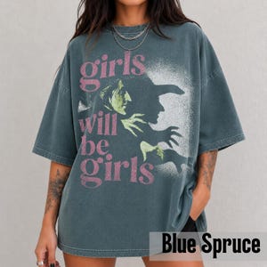 May include: A dark blue-grey oversized t-shirt featuring a graphic print of a green-skinned witch in profile. The text "girls will be girls" is printed in a distressed pink font over the image. The shirt has a crew neckline and short sleeves.