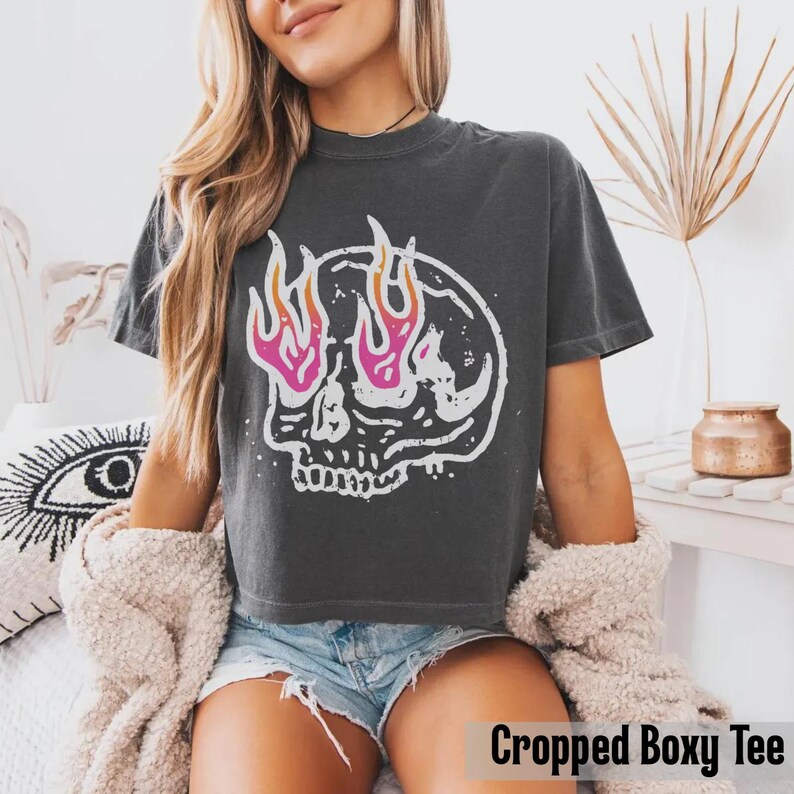 May include: Dark gray cropped boxy tee featuring a white skull graphic with pink and orange flames. The t-shirt has a crew neck and short sleeves. The text "Cropped Boxy Tee" is at the bottom of the image.