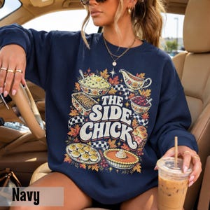 May include: A navy blue long-sleeve sweatshirt featuring a graphic print of various Thanksgiving side dishes, autumn leaves, and checkered patterns. The text reads THE SIDE CHICK in large white letters, surrounded by illustrations of food.