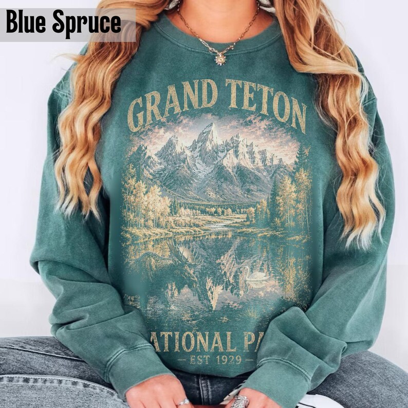 May include: Blue spruce-colored sweatshirt featuring a graphic of the Grand Teton National Park. The design includes a mountain range, trees, and a lake with the text "GRAND TETON NATIONAL PARK - EST 1929". The sweatshirt has long sleeves and a crew neck.