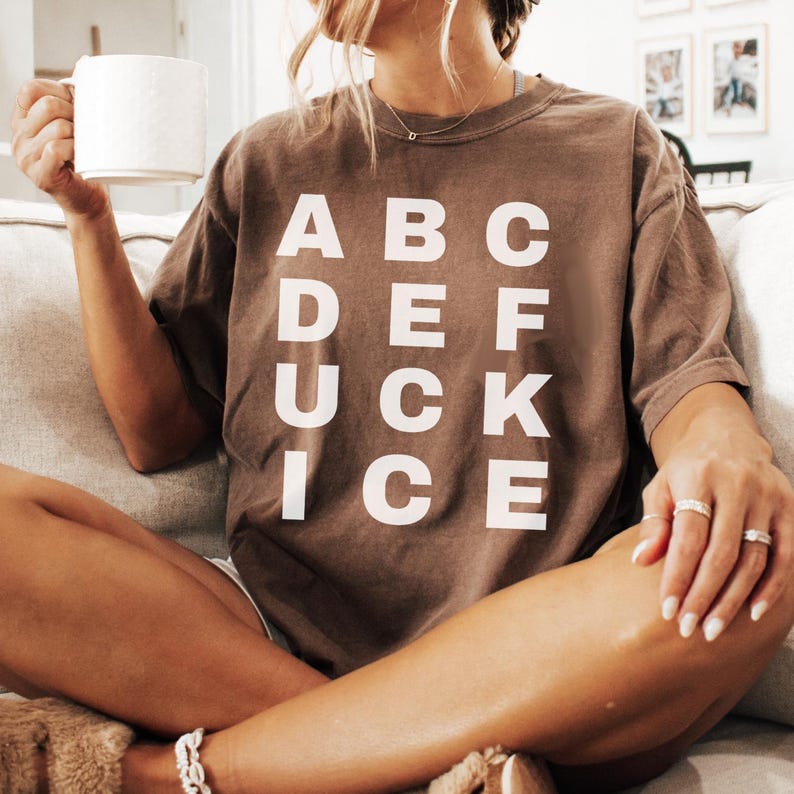 May include: A brown t-shirt with the text "ABC DEF UCK ICE" in white block letters. The shirt is worn by a person holding a white mug. The person is sitting on a couch.