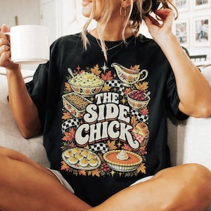 May include: A black short-sleeve t-shirt featuring a colorful graphic print of various Thanksgiving side dishes, including mashed potatoes, gravy, and pie. The text "THE SIDE CHICK" is printed in large, bold, cream-colored letters in the center.