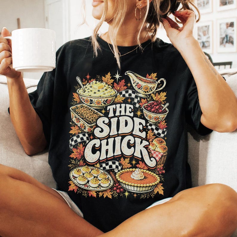 May include: A black short-sleeve t-shirt featuring a colorful graphic print of various Thanksgiving side dishes, including mashed potatoes, gravy, and pie. The text "THE SIDE CHICK" is printed in large, bold, cream-colored letters in the center.