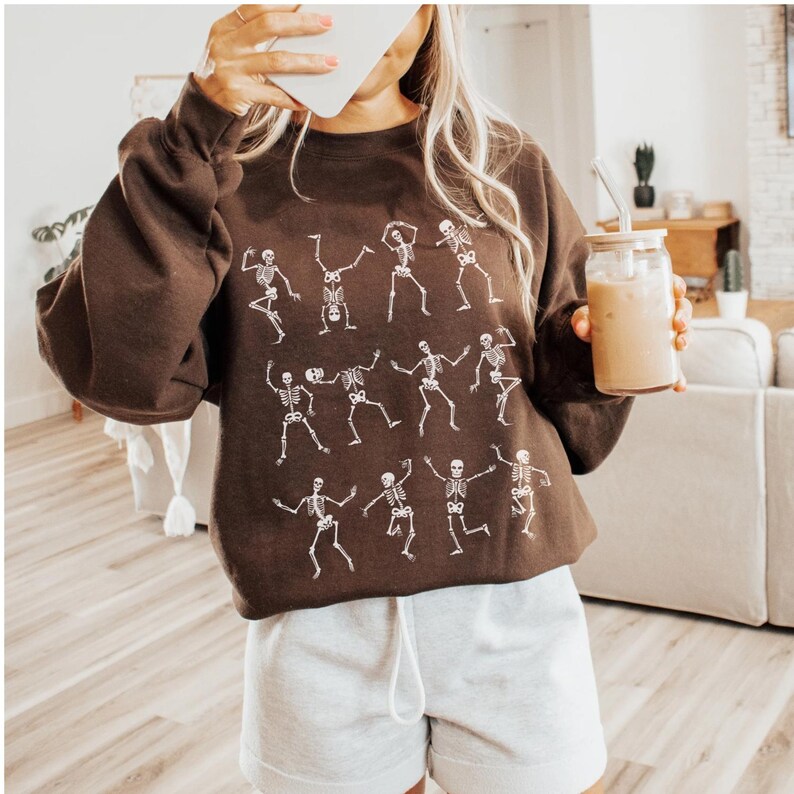 K&ouml;nnte beinhalten: Ein braunes Sweatshirt mit einem wei&szlig;en Skelett-Tanz-Design. Das lang&auml;rmlige Oberteil wird mit grauen Shorts kombiniert. Ein Glas mit Strohhalm und Getr&auml;nk wird in der Hand gehalten.