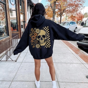 May include: Black hoodie featuring a skull design with flowers and a checkered pattern. The skull is in a golden color, and the flowers are in shades of orange and yellow. The hoodie is worn by a person on a sidewalk.