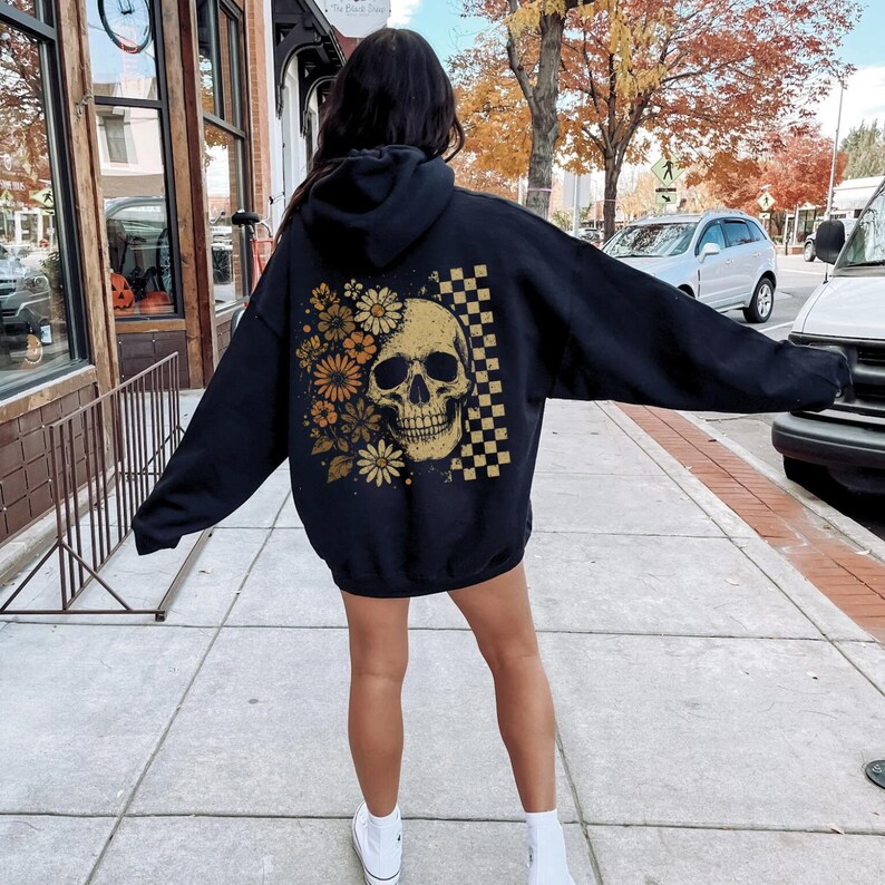 May include: Black hoodie featuring a skull design with flowers and a checkered pattern. The skull is in a golden color, and the flowers are in shades of orange and yellow. The hoodie is worn by a person on a sidewalk.