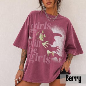May include: A berry-colored oversized t-shirt featuring a distressed white graphic of a witch in profile. The text "girls will be girls" is printed in a vintage-style font across the front. The shirt has a crew neckline and short sleeves.