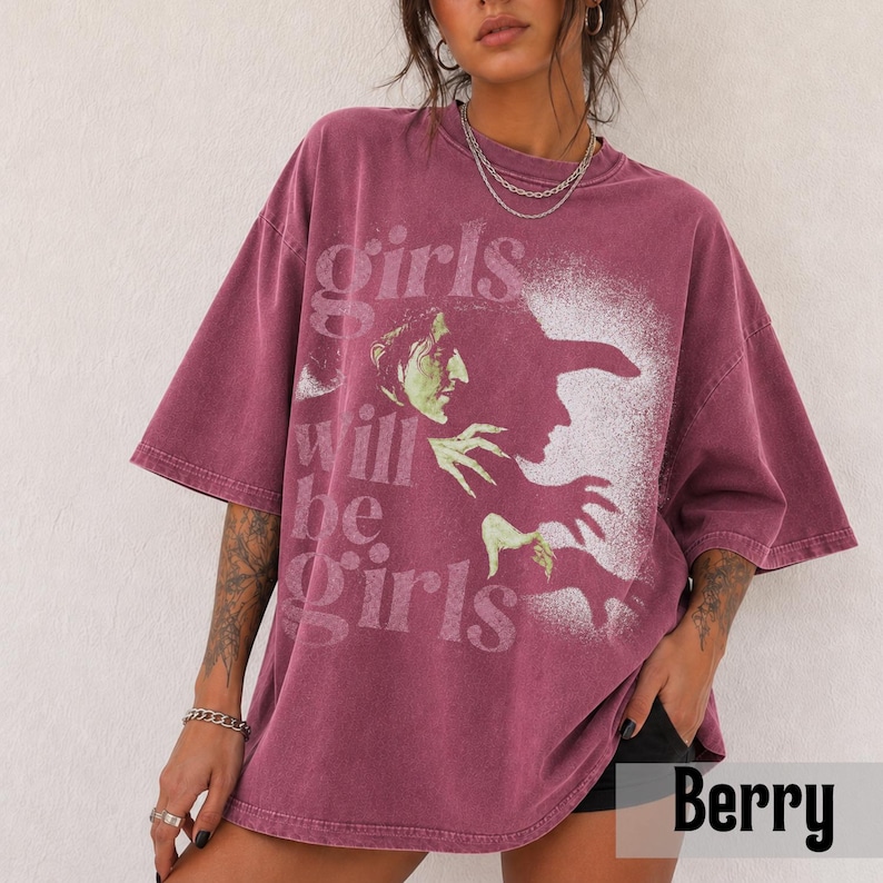 May include: A berry-colored oversized t-shirt featuring a distressed white graphic of a witch in profile. The text "girls will be girls" is printed in a vintage-style font across the front. The shirt has a crew neckline and short sleeves.