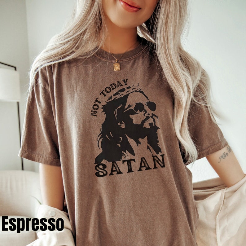 Pu&ograve; includere: Una maglietta marrone con una grafica di Ges&ugrave; che indossa occhiali da sole e una corona. Il testo "Not Today Satan" &egrave; stampato sulla maglietta.
