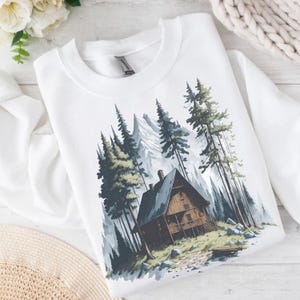May include: White sweatshirt featuring a watercolor illustration of a cabin nestled among tall pine trees and a mountain range. The cabin is brown with a dark gray roof, and the trees are green and gray.