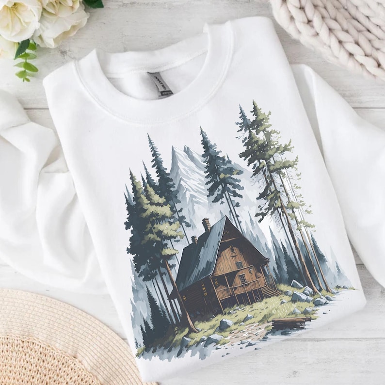 May include: White sweatshirt featuring a watercolor illustration of a cabin nestled among tall pine trees and a mountain range. The cabin is brown with a dark gray roof, and the trees are green and gray.