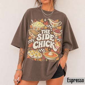 May include: A dark brown oversized t-shirt featuring a graphic print of various Thanksgiving side dishes, including mashed potatoes, gravy, and pie. The text reads THE SIDE CHICK in large white letters, surrounded by autumn leaves and checkered patterns.