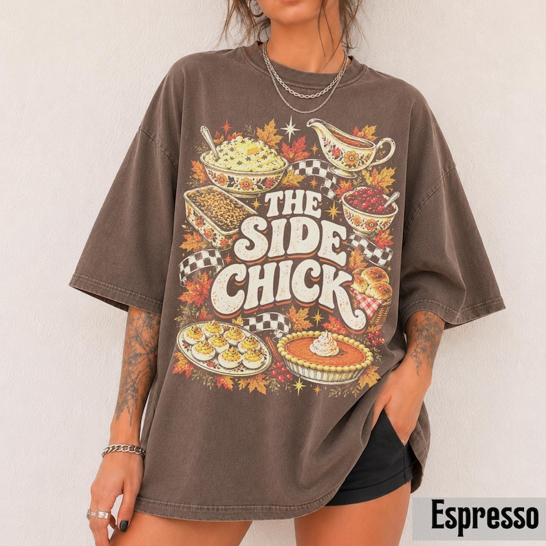 May include: A dark brown oversized t-shirt featuring a graphic print of various Thanksgiving side dishes, including mashed potatoes, gravy, and pie. The text reads THE SIDE CHICK in large white letters, surrounded by autumn leaves and checkered patterns.