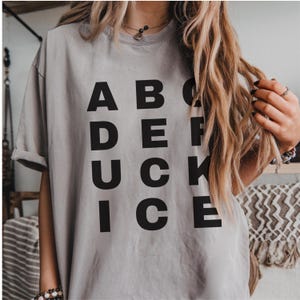 May include: A light gray t-shirt with the word "ADUCKICE" printed in large, black capital letters. The shirt has short sleeves and a relaxed fit. The model is wearing jewelry and has long, wavy blonde hair.