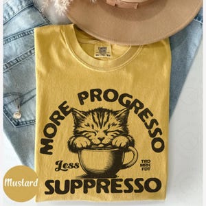 以下が含まれることがあります： マスタードイエローのTシャツに、コーヒーカップから飲んでいる猫の白黒のグラフィックがプリントされています。グラフィックには「More Progress Less Suppresso」というテキストと、猫の下に「TRD MRK FDT」というテキストが書かれています。