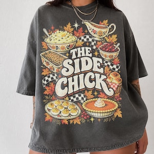 May include: A charcoal gray oversized t-shirt featuring a graphic print of various Thanksgiving side dishes, including mashed potatoes, gravy, stuffing, cranberry sauce, deviled eggs, and pumpkin pie, with the text THE SIDE CHICK.