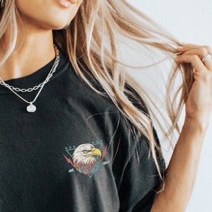 Op de afbeelding: Een zwart T-shirt met een afbeelding van een adelaar in een driehoek met sterren en de tekst "1947" op de voorkant. De adelaar is rood, wit en blauw.