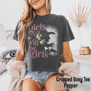 May include: A charcoal gray short-sleeve t-shirt with a boxy, cropped fit. The front features a graphic of a green-skinned witch in profile with a black hat, accompanied by the pink text "girls will be girls" printed in a stacked layout.