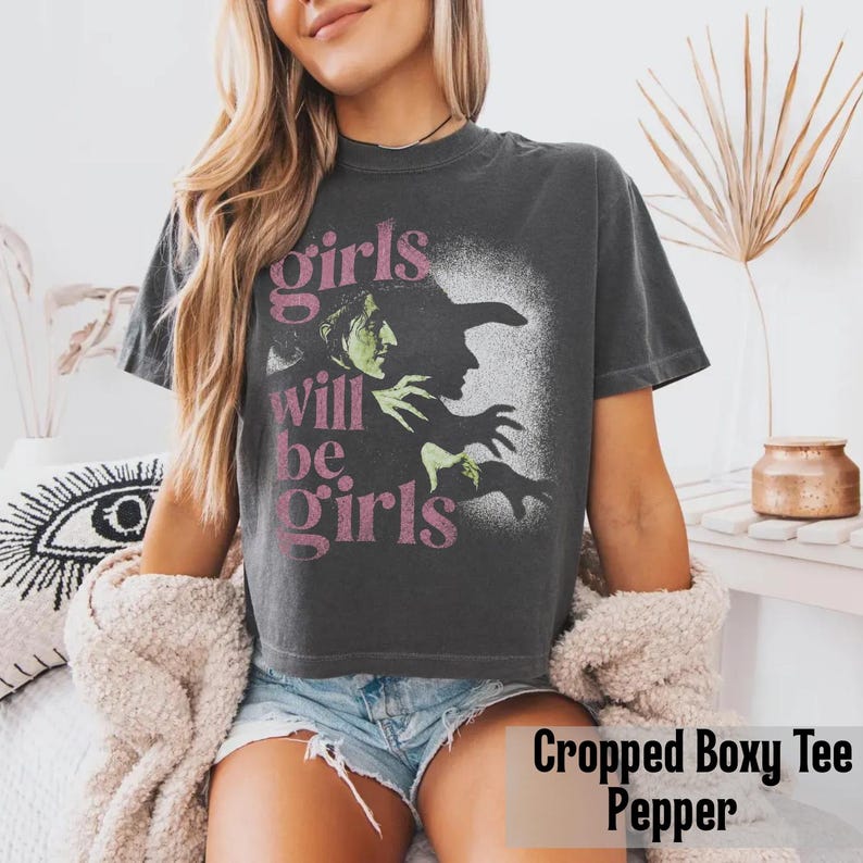 May include: A charcoal gray short-sleeve t-shirt with a boxy, cropped fit. The front features a graphic of a green-skinned witch in profile with a black hat, accompanied by the pink text "girls will be girls" printed in a stacked layout.