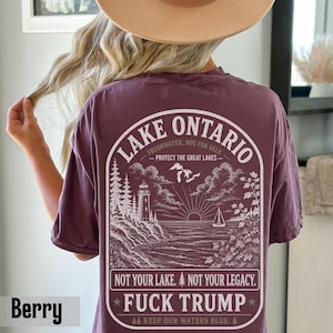 May include: A berry-colored t-shirt featuring a white graphic on the back. The design depicts a lighthouse, a sailboat, and Lake Ontario with the text: LAKE ONTARIO, FRESHWATER. NOT FOR SALE. PROTECT THE GREAT LAKES. NOT YOUR LAKE. NOT YOUR LEGACY. FUCK TRUMP.