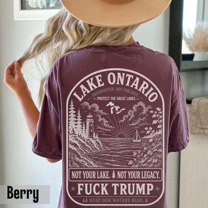 May include: A berry-colored t-shirt featuring a white graphic on the back. The design depicts a lighthouse, a sailboat, and Lake Ontario with the text: LAKE ONTARIO, FRESHWATER. NOT FOR SALE. PROTECT THE GREAT LAKES. NOT YOUR LAKE. NOT YOUR LEGACY. FUCK TRUMP.