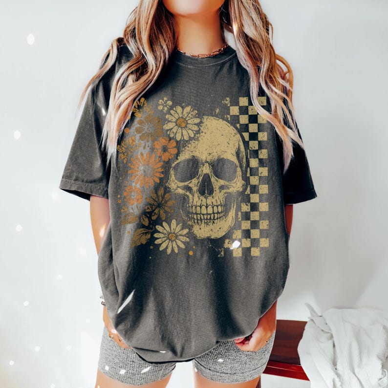May include: Dark gray t-shirt featuring a skull graphic with daisy flowers and a checkered pattern. The skull is in a vintage style with a distressed look. The shirt has short sleeves and a relaxed fit.